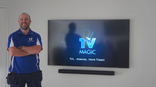 Technician with a freshly wall mounted TV, along with a wall mounted sound bar. TV Magic logo on the TV screen