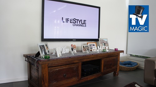 a large TV mounted on a wall above a cabinet with photo frames on it. TV Magic logo in top right corner