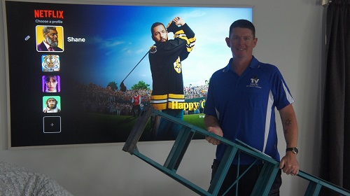 Technician - Brendan, holding a ladder in front of a wall mounted TV.