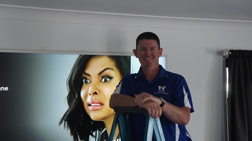 Technician - Brendan, in front of a large wall mounted TV.