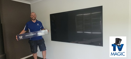 Technician - Vaughan, holding a TV Magic wall bracket, in front of a wall mounted TV. TV Magic logo in bottom right corner.