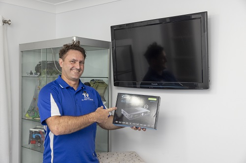 Technician holding a vast box, standing in front of a wall mounted TV.
