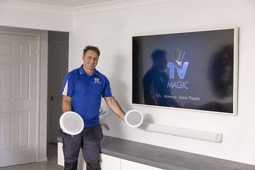 Technician holding two speakers in a living room with TV and soundbar.