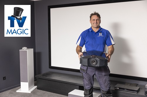 Technician standing in a home cinema holding a projector ready for installation. TV Magic logo in top left corner